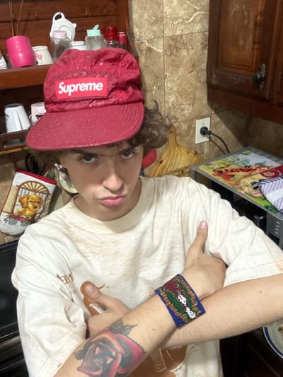 A person wearing a red Supreme cap and a beaded bracelet poses. The bracelet is blue with green, red, and white details. The person has a rose tattoo on their left forearm. They are wearing a light-colored t-shirt. A computer monitor sits nearby. The background includes kitchen cabinetry, tile flooring, and various household items.