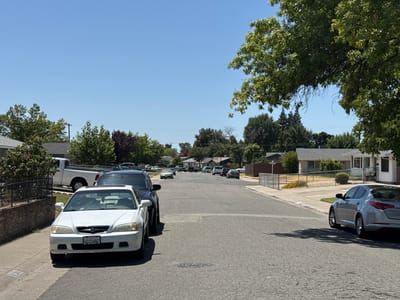 A sunlit, residential street in North Highlands, California, United States, is depicted on a clear, cloudless day. The scene is dominated by asphalt, with parked cars and houses lining both sides of the road. In the foreground, a white Acura sedan is parked on the left side of the street, its license plate reading "4GSS682". To its right, a dark-colored SUV is also parked. Further down the street, other vehicles are visible, including a white pickup truck on the left, and several sedans and SUVs parked or driving. Houses with pitched roofs and fences are situated behind the parked cars on the left, while on the right, a silver sedan is parked in front of a house with a light-colored exterior. Trees with lush green foliage are abundant, casting shadows on the street. The overall environment suggests a quiet suburban neighborhood.