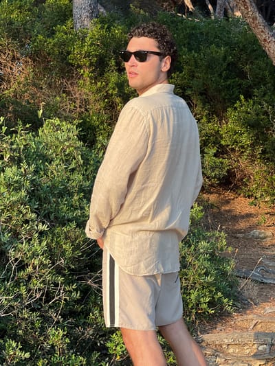 A man stands outdoors in a sunlit area with green foliage surrounding him. He wears a light beige linen shirt and matching shorts with a white and grey stripe detail. Dark sunglasses cover his eyes. His curly dark hair is visible. The background consists of a mix of green bushes, a tree trunk, and areas of direct sunlight.