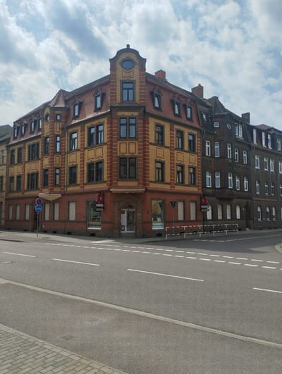 The image captures a street intersection in Bitterfeld-Wolfen, Germany, showcasing a distinctive corner building with a reddish-brown and yellow brick facade. This building has a corner entrance, multiple windows, and dormers on its red-tiled roof. To the right is a darker, older building with white-framed windows. Traffic signs are visible at the intersection, and a road with white dashed lines bisects the foreground. The sky is partly cloudy. There are no people visible.