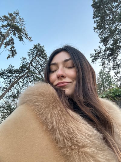 A low-angle selfie captures a woman with closed eyes, a serene smile, and long, dark hair. She is wearing a light beige coat with a fur collar. The background features trees with sparse foliage against a pale blue sky. The image is slightly rotated, giving a unique perspective. The woman's face is well-lit, and the composition emphasizes her relaxed expression.