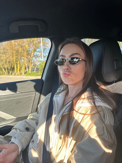 A woman wearing sunglasses and a light-colored jacket is sitting inside a car. The background visible through the car window is blurred and shows foliage and what appears to be the side of a road. The interior of the car, including the seats and a grab handle, are visible. There is no identifiable information to conclude the location is Nieporęt, Poland.