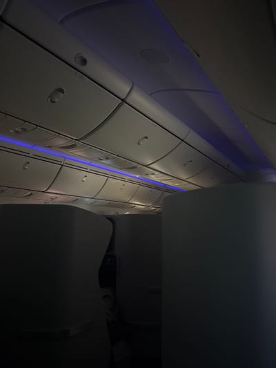 The image displays the interior of an aircraft cabin, located in Santo Domingo, Dominican Republic. The perspective is directed upwards, showcasing the ceiling and overhead compartments. The cabin is dimly illuminated by a prominent blue LED strip light running horizontally along the length of the ceiling, casting a blue ambient glow. Numerous closed, light-colored overhead luggage bins are visible, each equipped with individual passenger service units, including reading lights and call buttons. The ceiling itself is a curved, light-colored panel with additional recessed circular light fixtures. In the lower portion of the frame, the upper sections of at least three dark-colored, high-backed passenger seat structures, indicative of a premium cabin, are partially visible in shadow. No individuals, actions, or interactions are discernible within the frame.