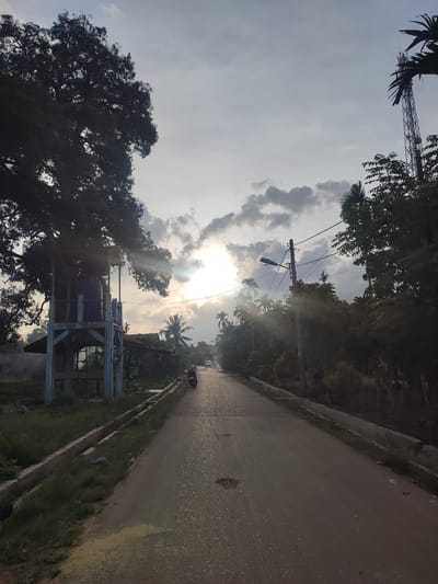 The image shows a street in Meunasahblangara, Indonesia, under a partly cloudy sky with the sun shining through. The asphalt road stretches into the distance, marked with a few potholes. A motorcyclist is traveling down the road. On the left side of the road, there is a water tower structure with a blue tank perched on top. A large tree provides shade over the structure. On both sides of the road, lush greenery and trees line the street. A utility pole with wires is visible on the right side. In the background, a cell tower rises above the trees. There are concrete drainage ditches along the road edges.
