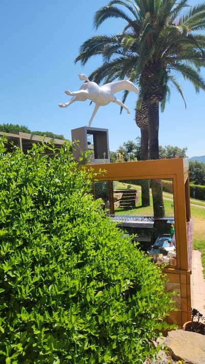 The image depicts a whimsical garden setting with a playful white statue of a dog in mid-leap, suspended in the air. The statue is positioned beneath a large palm tree, with its fronds reaching towards a clear blue sky. The scene is bathed in bright sunlight, suggesting it is daytime.
The garden itself appears to be well-maintained, with lush green foliage dominating the foreground and mid-ground. A prominent, thick hedge of vibrant green leaves runs across the lower portion of the image, obscuring some of the lower structures. Behind the hedge, a large, golden, rectangular frame is visible, acting as a portal to another part of the garden. Through this frame, one can glimpse manicured lawns, stone pathways, and structured hedges, creating a sense of depth and perspective.
To the left of the statue, a modern architectural element, possibly a balcony or a small building, is partially visible. Further into the background, more trees and greenery are scattered, hinting at a larger, expansive garden or park. The overall atmosphere is one of tranquility and amusement, enhanced by the bright, sunny weather and the serene natural surroundings. There are no people visible in the image. The location cues, such as the palm tree and the style of the garden, are consistent with a Mediterranean climate, and specifically with Lloret de Mar, Spain. There is no visible text in the image.