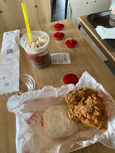 This overhead shot captures a casual meal at a KFC in Lhoksukon, Indonesia. The scene is set on a light-colored wooden table, suggesting an indoor dining area or perhaps an outdoor patio. The focus is on a KFC meal consisting of fried chicken, a serving of white rice, and a drink in a clear plastic cup with a yellow straw. Several small containers of red sauce, likely chili or ketchup, are arranged to the left of the meal. A small white packet, possibly a condiment or wet wipe, is also visible. A receipt with a QR code is lying at the bottom of the frame. The KFC packaging, with its distinctive red logo and branding, is prominent, wrapping the chicken and rice. The overall impression is one of a simple, satisfying meal, possibly during a daytime break. There are no people visible in the frame.