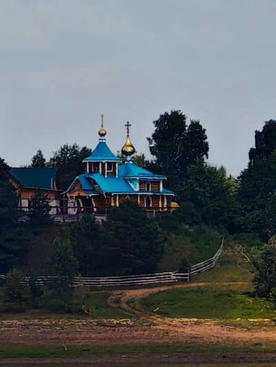 A wooden church with bright blue roofs and golden domes sits on a hill, surrounded by dense green trees. A white wooden fence lines a winding dirt path leading up the hill. The sky is overcast and gray. The image appears to be taken in Erkalovo, Russia.
