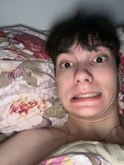 An individual with dark hair and fair skin is depicted in a close-up, head-and-shoulders photograph, positioned horizontally. The person is lying on a bed, partially covered by bedding featuring a floral pattern in shades of cream, red, and green on a light background. A thin, silver-colored chain necklace is visible around the neck. The individual's face displays an exaggerated expression, characterized by wide, staring eyes and a grimace, with the mouth open to reveal both upper and lower teeth, and the lips pulled back, causing visible wrinkles around the mouth, chin, and neck. The background directly behind the individual's head is a plain, light blue-gray surface. The specific location within the city of Zainsk, Russia, is not ascertainable from the visual evidence in the image.
