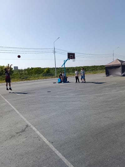 A group of young people are playing basketball on an outdoor court in Korsakov, Russia. The court is paved with asphalt and has white markings. A basketball hoop with a backboard and net is visible. One person is in mid-air, shooting the basketball. Several other individuals are standing or sitting on the court, observing the game. A large tent is set up to the right of the court. The background features trees and a clear blue sky.