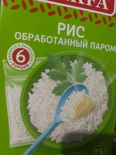 This is a close-up, overhead shot of a package of steamed rice, likely intended for sale in Kyrgyzstan given the Cyrillic text. The packaging is predominantly bright green with red oval shapes scattered across the left side. Dominating the center is a vibrant green circle filled with white, uncooked rice grains. A light blue spoon, tipped with a small piece of butter or margarine, rests on top of the rice. A sprig of fresh parsley is also placed on the rice, adding a touch of color and freshness.

To the left of the rice bowl, large white Cyrillic text reads "РИС" (Rice) and "ОБРАБОТАННЫЙ ПАРОМ" (Steamed). Below this, a circular emblem in red and white displays the number "6" and more Cyrillic text, which appears to relate to portion sizes or cooking instructions within sachets. On the right side of the package, more Cyrillic text is visible, including "25" and "Мин" (Minutes), suggesting cooking time, and "Пищевая ценность 100 г продукта" (Nutritional value per 100g product).

The lighting is bright and even, suggesting an indoor setting, possibly a kitchen or a retail display. There are no people visible in the image. The focus is entirely on the product packaging and its contents, highlighting the quality and preparation of the rice. The overall impression is one of freshness and simplicity, emphasizing the product's natural ingredients and ease of use.