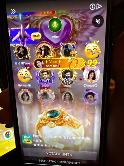 The image displays a close-up of a smartphone screen, held vertically. The phone is displaying a social media or chat application interface with numerous user avatars and text. The background of the app appears to be a purple, glittery design with a heart motif.
Prominently featured are several circular user profile pictures, some with animated elements. Text labels like "Vijay," "Veer," "Kavya," "Akash," "Rocky," and "Anjali" are visible, likely indicating user names. There are also icons such as a microphone, a mute symbol, and what appears to be a notification icon.
Towards the bottom of the screen, there is an app icon resembling a green square with a yellow smiley face. Adjacent to this icon, text reads "Hapi-Групповые голосовые чаты" (Hapi-Group Voice Chats), followed by star ratings and the word "УСТАНОВИТЬ" (Install). Below this, there is text that appears to be "БЕСПЛАТНО" (Free) and "РАЗВЛЕЧЕНИЯ" (Entertainment).
The overall impression is a snapshot of a mobile application interface, likely used for communication and social interaction. The image does not provide any visual information that would allow for the identification of the city of Małkowo, Poland, or any specific location within it.