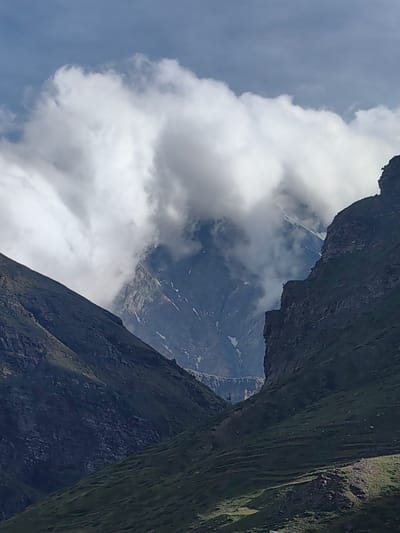 The image depicts a majestic mountain landscape, likely in the vicinity of Yamling, India. The foreground is dominated by steep, verdant slopes covered in grass and scattered rocks. These slopes converge to form a valley. In the midground, a rugged, rocky peak is partially obscured by dramatic, billowing white clouds that also frame the sky. Beyond this, a snow-capped mountain rises, its rugged texture visible through breaks in the clouds. The sky is a pale blue, with hints of grey in the clouds. The overall impression is one of a remote, wild, and breathtaking natural environment.