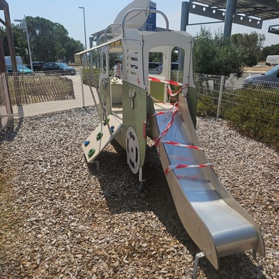 In the city of La Palme, France, a playground structure resembling a train or bus is cordoned off with red and white caution tape. The structure, painted green and white, features a climbing wall on the left with colorful grips, a steering wheel inside, and a silver slide on the right. The playground is covered in wood chips. In the background, a chain-link fence encloses a grassy area with trees and shrubs. Beyond the fence, cars are parked in a lot, and a solar panel canopy is visible. A sign with pictograms and text in French is mounted on a pole. A woman is partially visible inside the structure. The sky is clear and blue.