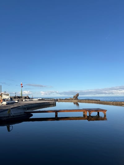 The image shows a pier extending into a body of calm, dark blue water under a bright, clear sky with a few wispy clouds. The pier is made of wooden planks and has a metal railing. To the right of the pier, an old, rusted metal structure, possibly a diving platform or a piece of abandoned infrastructure, stands partially submerged in the water. Its reflection is clearly visible on the smooth surface of the water.
Further down the pier, a navigational marker with red and white triangular flags and a green cone is visible, suggesting the area is near a harbor or a protected waterway. There are also some small boats docked along the pier. The scene appears to be set in the late morning or early afternoon, given the bright sunlight and the deep blue of the sky. The weather is clear and pleasant, indicative of a sunny day. The overall impression is one of a tranquil coastal environment, possibly a recreational area by the sea, with remnants of past industrial or recreational use. No people are visible in the image. The location context provided is Playa De Las Américas, Spain.