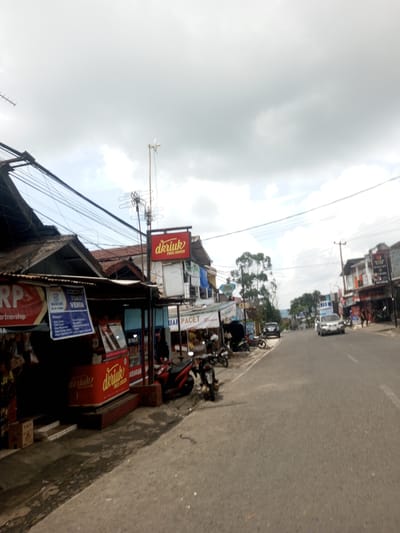 The image shows a street scene in Ban Phouviengnoy, Laos. The street is lined with small businesses, including a "dkriuk FRIED CHICKEN" restaurant. There are motorcycles parked along the sidewalk in front of the businesses. A man is standing in front of the "dkriuk" establishment. Cars are driving on the road. The sky is cloudy. Power lines are visible above the buildings.
