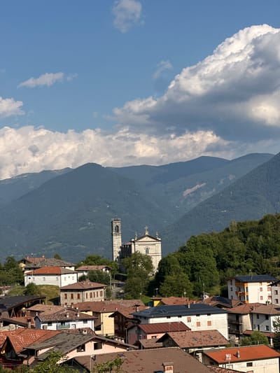 The image depicts a village nestled in the Italian Alps, specifically Ossimo Inferiore, Italy. The foreground is filled with a cluster of houses with terracotta roofs, showcasing varied architectural styles and colors, indicating a lived-in community. Dominating the mid-ground is a prominent church with a bell tower, its stone construction suggesting historical significance. Behind the village, lush green mountains rise, their slopes covered in dense forests. The sky above is a vibrant blue, dotted with fluffy white clouds that cast shadows on the landscape, suggesting a partly cloudy day. The overall scene conveys a sense of tranquility and natural beauty, typical of alpine villages. There are no people or animals visible in the image.
