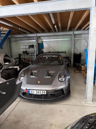A grey Porsche 911 GT3 RS is parked indoors within an industrial-style garage located in Meuspath, Germany. The vehicle is positioned facing the viewer, featuring a carbon fiber hood with twin air vents, a prominent black rear wing, and a Norwegian license plate "VJ 36530" with an attached red tow strap. A "MANTHEY" decal is affixed to the upper portion of the windshield. The garage structure consists of galvanized steel beams and columns, supporting an upper level with exposed wooden ceiling joists and fluorescent tube lighting. The walls are constructed of light-colored, corrugated metal panels. The concrete floor is visible. Ancillary objects within the garage include a stack of black tires on the left, a partially visible grey vehicle body on the far left, a shelving unit with various items in the background, a blue racing suit hanging on the rear wall, and engine components resting on a wooden pallet on the right. A carbon fiber panel is visible in the lower-left foreground, partially obscuring the view. No individuals are present in the scene, and no actions or interactions are depicted.