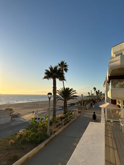 The image depicts an elevated view of a coastal scene in Manhattan Beach, United States, during late afternoon or early evening. The clear sky transitions from light blue overhead to a golden hue on the western horizon above the Pacific Ocean.

In the immediate right foreground, the side of a modern, multi-story residential building with light-colored walls and balconies featuring glass railings is visible. A white concrete railing marks the edge of the elevated vantage point.

A paved pedestrian pathway extends from the foreground into the midground, running parallel to the coastline. A single individual, dressed in a dark tank top and shorts, walks away from the viewer on this path. Further down the path, two other individuals are walking towards the viewer. The path is bordered on the left by a landscaped area containing various succulents, shrubs, and a low wooden fence, punctuated by decorative street lamps.

Beyond the pathway, a section of a paved road or parking area contains a parked white SUV. Adjacent to this road, the wide, sandy beach of Manhattan Beach extends to the ocean. Multiple small figures are visible on the beach, some near the water's edge and others near what appear to be beach volleyball nets.

The Pacific Ocean dominates the left side of the frame, showing gentle waves approaching the shore. Distant mountains or hills are faintly visible on the horizon, along with a large, indistinct vessel. Two tall palm trees and a shorter palm tree stand prominently in the mid-left, casting long shadows to the right. In the far distance along the coastline to the right, industrial structures, including tall stacks, are discernible.