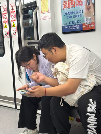 Two individuals, a woman and a man, are seated on a bench inside a subway car. The woman, on the left, is wearing a light purple top and black pants, with dark hair. She is holding a smartphone and looking at it. The man, on the right, is wearing a white t-shirt and black pants with white lettering. He is wearing glasses and has short dark hair. He has an arm around the woman and is also looking at the phone. They are seated next to each other. To their left, a subway door is visible, adorned with various warning signs including "No Smoking," "No Loud Noise," "Beware of Closing Doors," and "Mind the Gap." To their right, a large advertisement is displayed on the wall, featuring Chinese text and a QR code. The advertisement appears to be for a dental clinic. The subway car interior is visible, with a metal pole to the left and the floor is a light gray color. The overall lighting is bright, typical of public transportation.