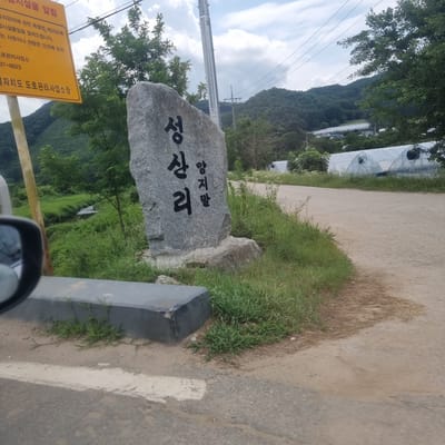 The image depicts a stone monument with Korean characters carved into it, standing amidst lush green grass and vegetation. The monument appears to be located in a rural setting, possibly in Anheung-myeon, South Korea, given the presence of greenhouses in the background and a dirt road. The sky is partly cloudy, with a clear view of distant hills covered in trees. A yellow sign with Korean text is partially visible on the left side of the image, suggesting it might be a place of interest or a directional marker. The overall scene conveys a sense of tranquility and nature.