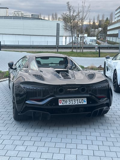 The image shows the rear of a black McLaren Artura, with the Swiss license plate "ZH-313140". The car is parked on a paved surface, possibly a parking lot. A partially visible white car is present in the background. The "McLaren Artura" branding is visible on the back of the vehicle. The location is indicated by the license plate, which designates Zurich (ZH), Switzerland. The presence of the "McLaren Zürich" dealership further suggests the location. The scene includes buildings, trees, and a cloudy sky. Specific city location is Glattbrugg, Switzerland.