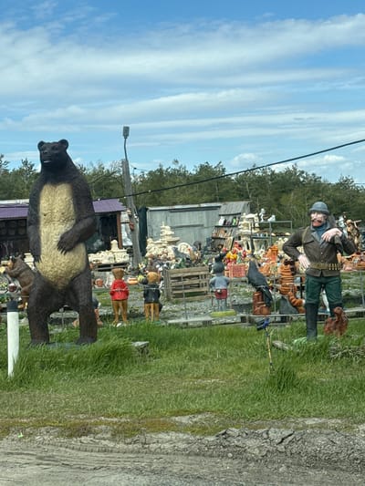 The image shows a cluttered outdoor space filled with various statues and decorative objects, potentially a roadside attraction or garden display in Umet, Russia. The environment consists of a mix of grass, gravel, and a cracked asphalt surface. In the foreground, a large bear statue dominates the scene. Behind the bear, a diverse assortment of other statues and objects are visible, including a man in military garb, a tiger, and numerous smaller figurines. A simple building with a corrugated metal roof is in the background. Utility poles and overhead wires are visible against a blue sky with clouds. The scene appears haphazard, with objects densely packed together.
