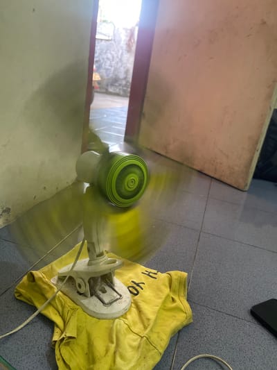 In Anggaswangi, Indonesia, a white electric fan with a green grille is clipped onto a yellow t-shirt lying on a tiled floor. The fan is in operation, indicated by the blurred motion of its blades, which creates a yellow glow around the device. The background reveals a dimly lit interior with a doorway leading to an outdoor area. The walls are textured and appear weathered. A large, light-colored rectangular object, possibly a door or a piece of furniture, is visible on the right side of the image. A dark rectangular object, likely a smartphone, is on the floor to the right of the t-shirt. The scene suggests a domestic setting where the fan is being used to cool the area.