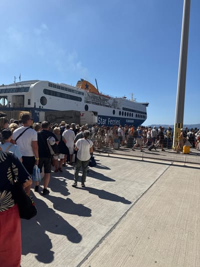A crowd of people are lined up on a concrete pier, waiting to board a large ferryboat named "Star Ferries". The ferry is white with blue accents and has "Star Ferries" printed in large blue letters on its side. The boat is docked at a port in the city of Thira Municipal Unit, Greece, with a clear blue sky overhead. Many people in the crowd are carrying luggage. The scene suggests a common mode of transportation or tourism within the area.