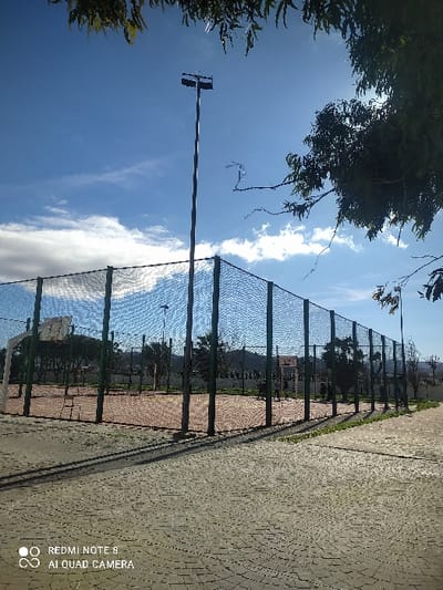 The image depicts an outdoor sports facility, specifically a basketball court, located in Tétouan, Morocco, under a bright, partly cloudy blue sky.
In the foreground, a grey, patterned paved surface extends, with long, dark shadows from fence posts and overhead foliage cast upon it, indicating strong sunlight. The central subject is a rectangular basketball court with a reddish-brown surface, partially visible court lines, and a basketball hoop structure on the left side, including a backboard and net. The court is enclosed by a tall fence composed of dark netting stretched between green-colored vertical posts.
Several indistinct human figures are visible within the court, suggesting individuals are present and possibly engaged in activity. Behind the court fence, a line of green trees and shrubs is visible, with distant hills and faint structural outlines beyond them.
Two tall, grey metal lighting poles are present: one prominently centered behind the court fence with two horizontal light fixtures, and another shorter pole partially visible on the far right. The upper portion of the frame is dominated by the blue sky and scattered white clouds, with out-of-focus green leaves and branches from an overhead tree encroaching from the top and upper-right. A "REDMI NOTE 8 AI QUAD CAMERA" watermark is present in the lower-left corner.