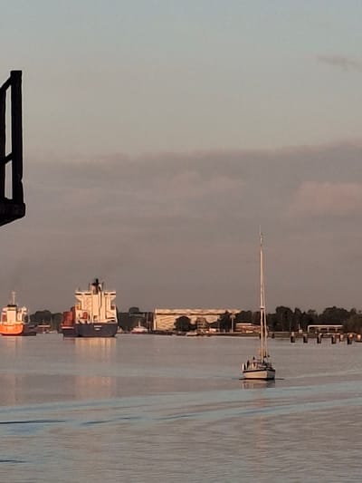 The image shows a waterway in Brunsbüttel, Germany, at dusk. Several vessels, including cargo ships and a sailboat, are visible along the waterway. The shoreline features buildings and trees. A black structure is in the foreground, possibly part of a pier or dock. The sky is overcast.