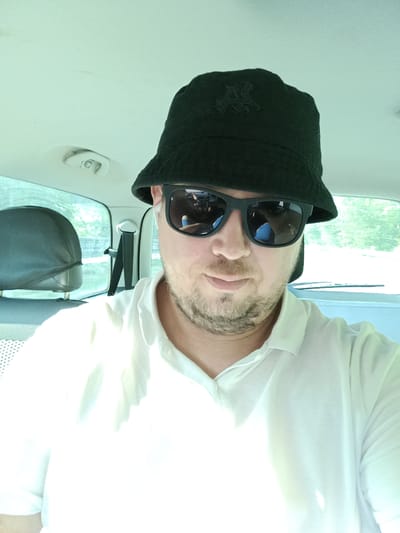 This is a selfie taken by a man sitting in the passenger seat of a car, likely in Ostředek, Czechia. He is wearing a black bucket hat with an embroidered logo and black sunglasses. He is also wearing a white polo shirt. The photo is taken during the day, with bright, diffused light, suggesting it might be a sunny day. The background shows the interior of the car, including a headrest of the back seat and a seatbelt. Through the windows, trees and greenery are visible, indicating an outdoor, possibly rural or suburban setting. The man's expression is neutral, and he appears relaxed. There is no visible text in the image.
