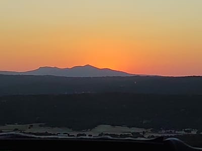 The image shows a wide landscape view of a mountain range at sunset. The sky transitions from a pale yellow at the top to a vibrant orange as it nears the horizon. The sun, partially obscured by the mountains, is a bright orange disc. A dark, silhouetted mountain range runs horizontally across the middle of the image, with subtle variations in elevation creating a jagged skyline. Below the mountains, a dark, densely forested area fills the lower middle section of the frame. In the foreground, the lower edge of the image is occupied by a dark, out-of-focus area, possibly representing the edge of a vehicle or a foreground structure. The lighting suggests that the photograph was taken during the golden hour, just before or during sunset, casting a warm glow over the scene. The image appears to be taken from a high vantage point, overlooking the landscape.