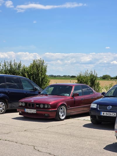 The image, captured in Akatsievo, Bulgaria, displays a sunny outdoor scene with a clear blue sky dotted with fluffy white clouds. In the foreground, a row of cars is parked on a cracked asphalt surface. The primary subject is a maroon BMW sedan with its front prominently visible. The car has a license plate that reads "CB 0989 KM" and displays the text "Auto Bavaria." To the left of the BMW, a dark blue SUV is partially visible. To the right of the BMW, the front of a dark blue Volkswagen sedan is seen, with its license plate showing "M 8436 CA." Beyond the cars, a line of lush green trees separates the parking area from a field of golden crops, suggesting a rural or semi-rural setting. The overall impression is one of a static, parked collection of vehicles under a bright, summery sky.