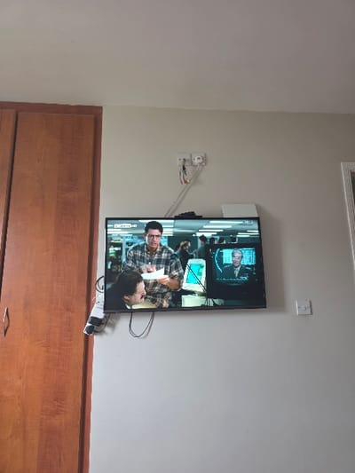 The image displays an indoor scene featuring a wall-mounted black flat-screen television and a wooden wardrobe. The specific location within Tullamore, Ireland, is not discernible from the visual evidence. The television screen presents a scene with two male subjects: one male in a plaid shirt and eyeglasses stands, holding a white document, while another male with dark hair and a mustache looks up at him. They are positioned within an environment that includes additional blurred figures and what appear to be older computer monitors. A smaller picture-in-picture display on the television shows an older male in a suit. The 'SKY NEWS HD' logo is visible in the top-left corner of the main screen. Above the television, two white double electrical outlets are affixed to a light beige wall, with connected red, white, and yellow cables. A white power strip with multiple black cables hangs to the left of the television, partially obscured by the wardrobe. A single white electrical outlet is mounted on the wall to the right of the television. A tall, two-door wooden wardrobe with a warm brown finish and a silver-colored handle occupies the left side of the frame. A white ceiling is visible at the top of the image, and a fragment of a white doorframe is present on the far right edge.