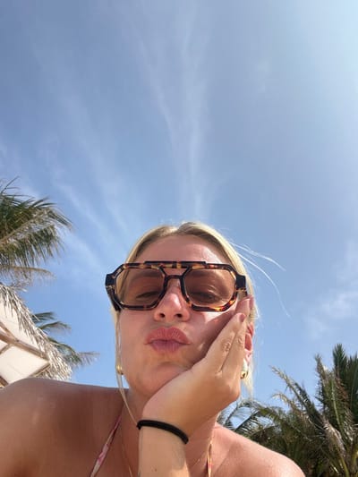 A woman wearing tortoise-shell sunglasses stands with her hand over her forehead. She pouts her lips towards the camera. Above her is a bright blue sky with light clouds and fronds of palm trees. She wears a black bracelet on her right wrist. A sliver of a white structure is visible at the bottom of the photo.