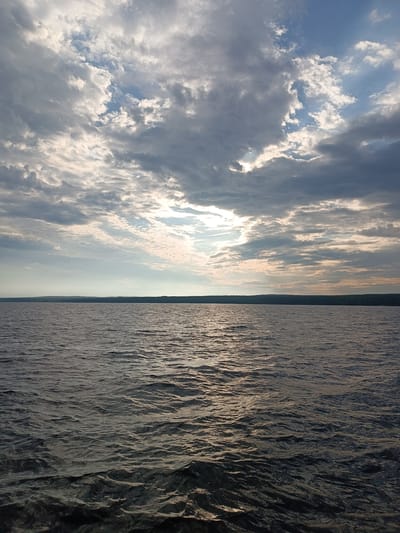 The image shows a wide expanse of rippling water, likely a reservoir or large river, under a dramatic sky filled with a mix of dark and light clouds. Sunlight breaks through in some areas, casting a soft glow on the water's surface and creating reflections. The horizon is lined with a dark, silhouetted forest, suggesting a natural, undeveloped landscape. The water appears calm but with a slight chop, indicating a gentle breeze. There are no people or man-made structures clearly visible in the foreground or middle ground. The scene evokes a sense of tranquility and natural beauty. Given the information provided, the image could potentially be of the Kama River or the Votkinsk Reservoir near Chaikovsky, Russia, an area known for its large bodies of water and surrounding forests.