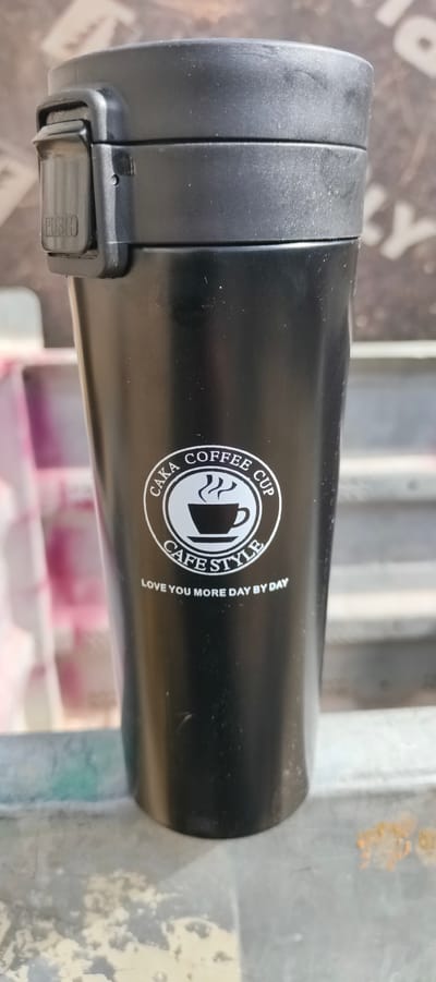 A black thermos cup with a white circular logo is the primary subject, presented in a close-up shot. The logo features a steaming coffee cup design and text. The top of the thermos has a black lid with a silver "PUSH" button clearly visible. The background is blurred, showing a mix of muted colors, including grays and hints of pink, suggesting an outdoor or possibly indoor setting with some distance. The lighting appears natural, indicating daytime. The cup is resting on a textured surface that seems like a bench or ledge, with some specks of dirt or debris.
Visible text on the cup reads:
- "PUSH" on the lid mechanism.
- "CAKA COFFEE CUP" encircling the top of the logo.
- "CAFE STYLE" below the coffee cup graphic.
- "LOVE YOU MORE DAY BY DAY" below "CAFE STYLE".