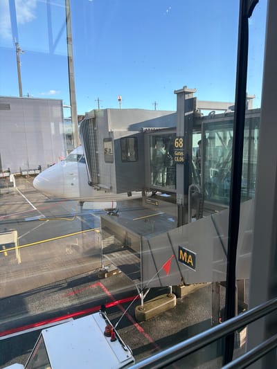 This image depicts a view from inside an airport terminal building, looking out through a window at an airplane parked at a gate. The airplane's nose and front fuselage are visible, connected to a jet bridge. The jet bridge is a gray, enclosed walkway with large glass panels, allowing views of people inside. A sign on the jet bridge reads "68 Gates C6, D6". Another sign shows "MA". The ground area around the airplane and jet bridge is paved and marked with yellow and red lines. A white airport vehicle is partially visible in the foreground. The sky is clear and blue with a few scattered clouds. The scene is outdoors at an airport, likely Oslo Airport, Gardermoen, Norway, as indicated by the "MA" possibly representing "Main Airfield" or a specific airline/airline code used at that airport. The lighting suggests it is daytime.