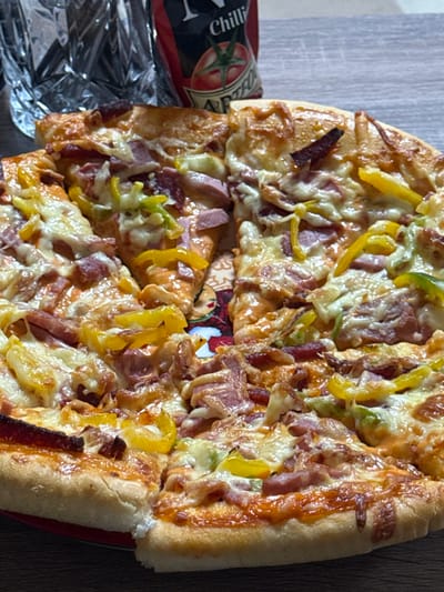 The image presents a close-up, slightly elevated view of a large, round pizza, sliced into multiple portions, resting on a patterned plate with red and brown decorative elements. The pizza features a thick, golden-brown crust and is topped with melted, bubbly cheese, numerous pieces of pinkish-red processed meat (consistent with ham or cured sausage), and slices of yellow and some green bell peppers. In the blurred background, a clear, textured glass object is partially visible on the left. To its right, a red bottle with a white label showing the word "Chilli" and a tomato graphic is also out of focus. The items are situated on a dark, likely wooden, tabletop. No individuals or active interactions are visible in the scene, which is located in Taperakan, Armenia.