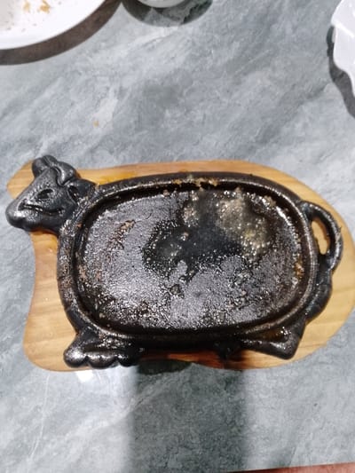 The scene is set on a mottled grey, textured tabletop, presumably in Thôn 8, Vietnam. A dark, cast iron sizzling plate, shaped like a stylized cow, occupies the central foreground. The plate features an oval-shaped well containing dark, glistening residue, likely oil and small fragments of cooked food. This cast iron plate rests on a light brown, contoured wooden base that mirrors its shape. In the upper left background, a portion of a white ceramic plate is visible, containing a few light-colored crumbs or food particles. Another indistinct white object is partially visible in the upper right background. No people are present. The lighting is direct, creating reflections on the greasy surface of the sizzling plate.