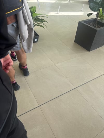 The image shows a person sitting on a tiled floor next to two potted plants. The person is wearing shorts, black shoes, and pink socks, and is wearing a black jacket. The location is inferred to be in Mellieħa, Malta.