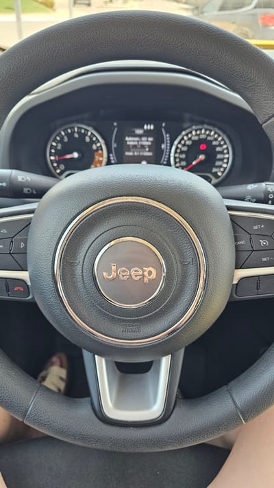 The image shows a close-up view of a Jeep steering wheel from the driver's perspective. The steering wheel is black and textured. The Jeep logo, a circular emblem with the word "Jeep" in raised gold lettering, is prominently displayed in the center. The dashboard is partially visible, with a digital display showing some information, including what appears to be a temperature reading of 400 degrees (likely a malfunction or misinterpretation of data). There are also two circular gauges, likely the speedometer and tachometer. On the left side of the steering wheel, there are buttons for controlling the vehicle's functions, including cruise control. On the right side, buttons labeled "SET+", "CANC", and "SET-" are visible, along with other control buttons. The bottom center of the steering wheel has the "SRS AIRBAG" text. Below the steering wheel, a person's legs and feet are visible, wearing sandals with a beige and white striped pattern. The person is wearing black pants. The overall environment appears to be inside a car, possibly parked or in motion, with a blurred outdoor scene visible through the windshield. The image does not provide any explicit visual clues to its specific location in María De Huerva, Spain.
