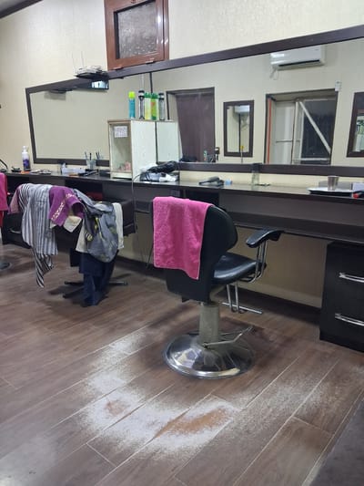 The image shows a dimly lit interior of what appears to be a salon or barbershop. The scene is dominated by a large mirror that reflects the room, making it difficult to discern the exact layout. A black salon chair, partially covered with a pink towel, is the central object. The floor is made of wooden planks, and there is a noticeable scattering of white powder on the floor near the chair.
On the shelves visible in the mirror's reflection, there are various bottles of toiletries, brushes, and other salon tools. A black hairdryer is also visible. The overall impression is of a workspace that may have recently been used or is in the process of being cleaned. There are no people visible in the image.
Some text is visible on a small sign in one of the reflected areas: "EHSON QUTISI." The lighting is artificial and casts shadows, suggesting it is likely not daytime or the blinds are drawn. The setting appears functional and somewhat cluttered, typical of a service establishment. The presence of the white powder suggests a possible cleaning or styling product residue. The overall mood is neutral and focuses on the objects and the setting itself.