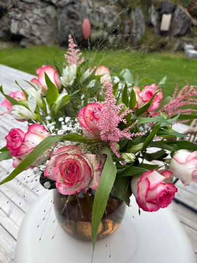 A bouquet of pink and white roses is displayed in a brown glass vase on a white table. The table is situated outdoors, with a wooden deck visible beneath it. In the background, a grassy area leads to a rocky outcrop. The scene is likely in Lommedalen, Norway, given the context. The lighting suggests it is daytime. There are no people or animals present in the image.