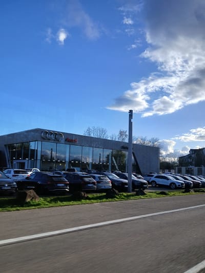 The image displays the Audi Zentrum Homburg Saar GmbH dealership, located at Daimlerstraße 1, 66424 Homburg, Germany. The dealership building is a modern, two-story structure with a grey, horizontally ribbed facade and an extensive glass-paneled front that reflects the sky and clouds. The prominent Audi rings logo and the red "Audi" text are mounted on the upper facade.

In front of the dealership, a large inventory of Audi vehicles, primarily new and used SUVs and sedans in various dark and light colors, are parked in multiple organized rows on an asphalt surface. A low, green grass median containing several dark, irregular rocks separates the dealership's parking area from the foreground. The foreground consists of an asphalt road with a solid white line, exhibiting motion blur consistent with a photograph taken from a moving vehicle. A tall, slender grey pole, possibly a light fixture or surveillance mast, stands vertically within the parking lot.

The sky overhead is predominantly blue with scattered white cumulus clouds, and larger, darker grey cloud formations are visible towards the upper right. Bare deciduous trees are visible behind and to the right of the dealership building, suggesting a late autumn or winter season. A multi-story building, possibly residential or commercial, with dark windows, is partially visible in the far right background beyond the trees. No individuals are distinctly visible within the frame, and no specific actions or interactions are depicted.