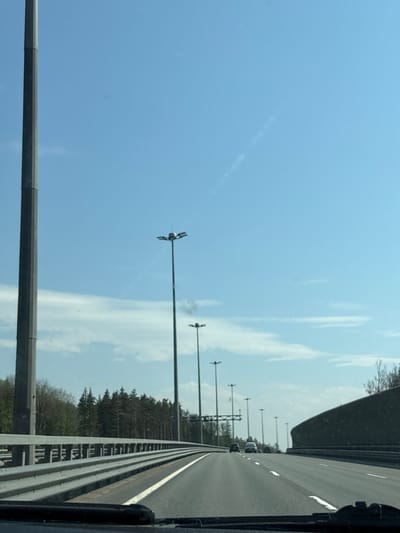 The image shows a highway scene in Pesochny, Russia, viewed from the perspective of a vehicle moving along the road. The sky is blue with wispy clouds. Rows of highway light poles line the road. A barrier runs along the edge of the highway, separating the road from a forested area. Cars can be seen in the distance.