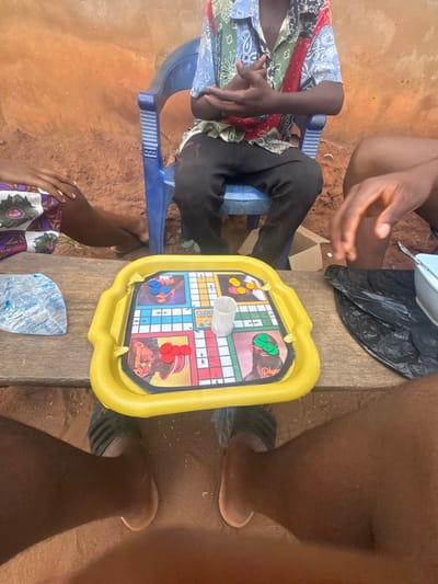 Four people gather around a yellow board game, likely Ludo, placed on a wooden surface. The game features faces of what appears to be musicians. A person wearing a colorful shirt sits on a blue plastic chair, while another wears a patterned skirt. A white dice cup sits in the center of the board, surrounded by game pieces. The scene is shot at an awkward angle, possibly from above, and includes partially visible legs and feet of the individuals playing.
