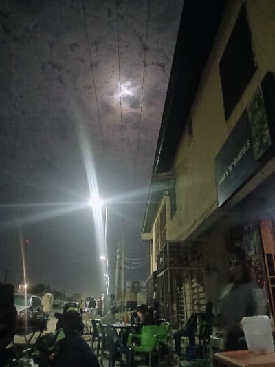 This image depicts a nighttime street scene in Mumbwa, Zambia. The sky is dark and cloudy, revealing a partially obscured moon and multiple overhead power lines. Artificial light sources, including streetlights, illuminate the scene; a prominent streetlight in the mid-left creates a significant lens flare.
On the right, a light-colored, multi-story building features dark rectangular windows and a sign reading "TIMES N' MOVIES" on its facade. A metal cage structure is visible at the building's ground level. Along the street, utility poles with power lines extend into the background.
In the foreground and midground, an outdoor seating area is present, furnished with small tables and an assortment of plastic chairs, some green, some dark. Approximately 10-15 individuals are dispersed throughout the scene. Several people are seated at tables, engaged in various activities such as conversing or using mobile devices. A person in a light-colored shirt stands on the far right, partially obscured by a white container resting on a wooden surface. Further down the street, additional individuals and at least one light-colored vehicle are visible.