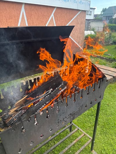 The image captures an outdoor scene featuring a burning barbecue grill. The grill itself is made of dark metal and is filled with burning logs that produce vibrant orange flames and some wisps of smoke. The grill is positioned on a grassy area, with a hint of a wooden bench visible in the background. Beyond the immediate grilling area, there are buildings and lush green trees, suggesting a rural or suburban setting. The overall impression is that of a warm, sunny day, ideal for an outdoor gathering or cooking activity. There is no text visible in the image.