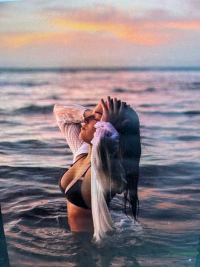 The image shows a woman standing in the ocean. She is wearing a black bikini and a white sheer shirt. Her hands are raised to her head, and her eyes are closed. The ocean is calm, and the sky is a soft pink and orange, indicating it is either sunrise or sunset. There are no visible landmarks that would suggest a specific location in Navahrudak, Belarus.